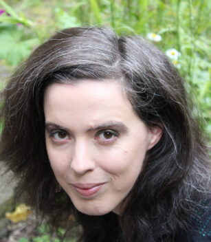 Eine Frau mit langen, dunkelbraunen Haaren und einigen grauen Strähnen blickt in die Kamera; im Hintergrund sind im Freien grüne Pflanzen und Gänseblümchen zu sehen.