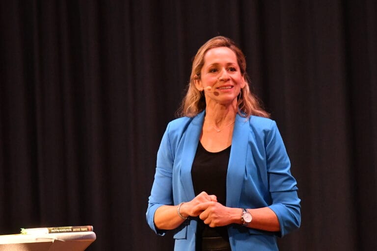 Eine Frau in blauem Blazer und schwarzem Hemd steht mit einem Mikrofon-Headset auf der Bühne und spricht vor einem dunklen Vorhang, neben ihr ein Tisch mit Büchern.
