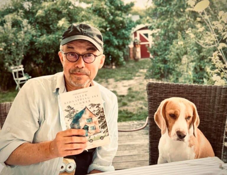 Ein Mann mit Brille und Mütze sitzt an einem Tisch im Freien und hält ein Buch in der Hand. Ein Beagle-Hund sitzt neben ihm. Im Hintergrund sind Grünpflanzen und ein roter Schuppen zu sehen.