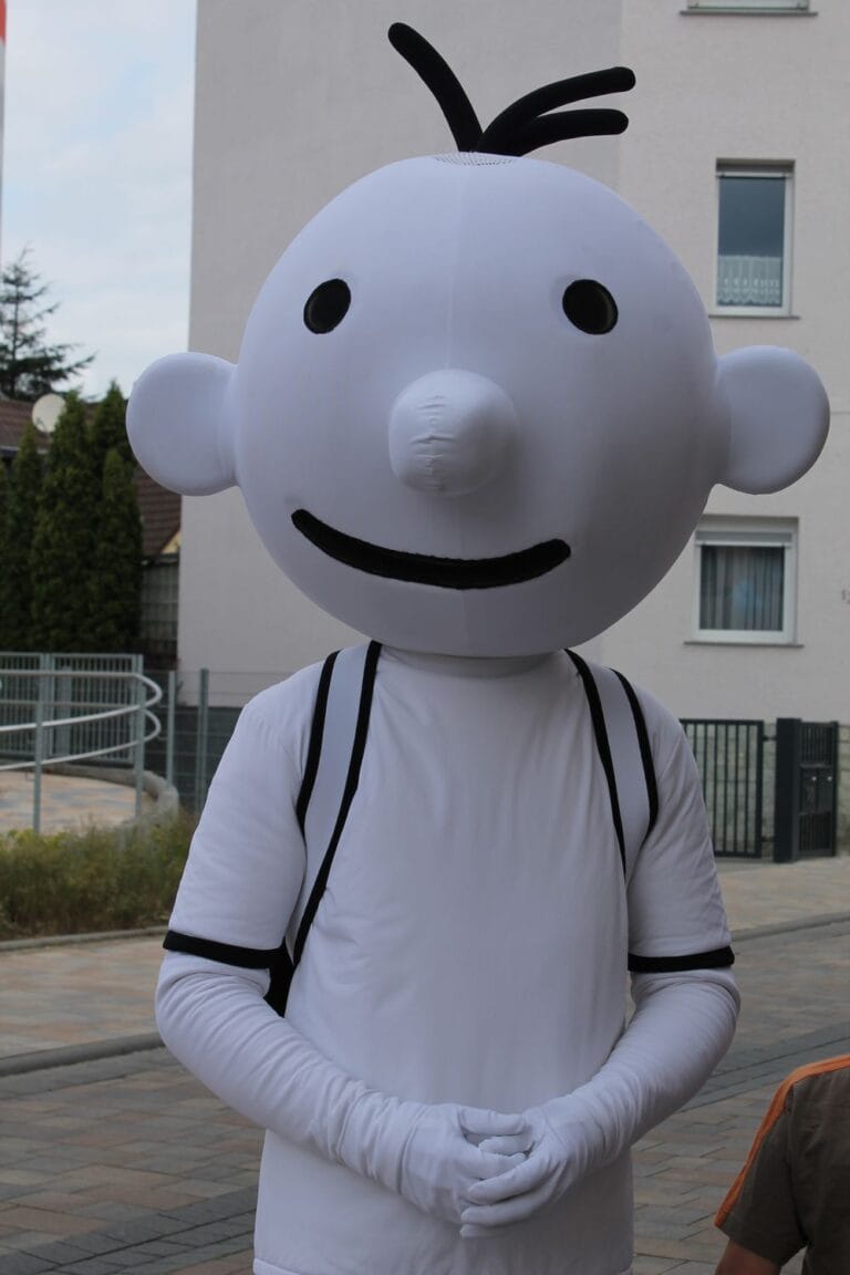 Person, die ein großes weißes Zeichentrickfigurenkostüm mit rundem Kopf, schwarzen Augen und einem schwarzen Lächeln trägt und im Freien auf einer gepflasterten Straße steht.