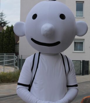 Person, die ein großes weißes Zeichentrickfigurenkostüm mit rundem Kopf, schwarzen Augen und einem schwarzen Lächeln trägt und im Freien auf einer gepflasterten Straße steht. {{brizy_dc_image_alt entityId=