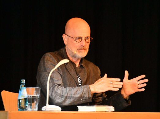 Berkel 1 (17) Ein glatzköpfiger Mann mit Brille und Bart spricht, während er mit den Händen gestikuliert. Er sitzt an einem Tisch mit einer Lampe, einer Wasserflasche, einem Glas und einem Buch, vor einem dunklen Hintergrund.