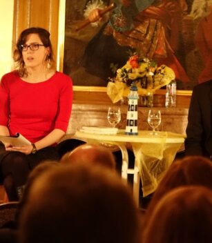 Eine Frau in einem roten Oberteil und ein Mann in einem dunklen Anzug sitzen an einem kleinen Tisch auf der Bühne, dem Publikum zugewandt in einem warm beleuchteten Raum mit einem Gemälde und einem Blumenarrangement hinter ihnen. {{brizy_dc_image_alt entityId=