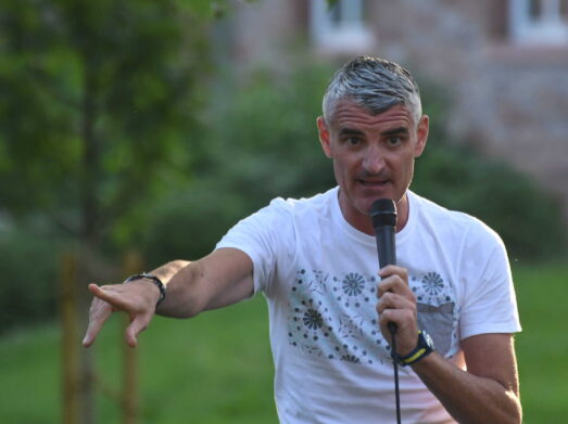DSC_6473 Ein Mann mit kurzen grauen Haaren in einem weißen T-Shirt hält ein Mikrofon und gestikuliert mit der anderen Hand, während er im Freien spricht. Ein unscharfer grüner Hintergrund ist sichtbar.