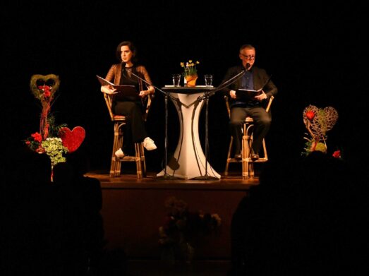 Zwei Personen sitzen auf einer Bühne und lesen Bücher. Zwischen ihnen steht ein kleiner runder Tisch mit Wassergläsern und einem Blumenarrangement. Die Bühne ist mit dekorativen herzförmigen Aufstellern geschmückt.