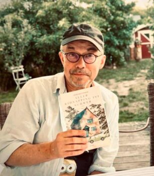Ein Mann mit Brille und Mütze sitzt an einem Tisch im Freien und hält ein Buch in der Hand. Ein Beagle-Hund sitzt neben ihm. Im Hintergrund sind Grünpflanzen und ein roter Schuppen zu sehen. {{brizy_dc_image_alt entityId=